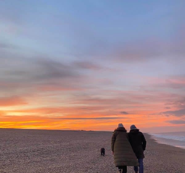 A couple walk their dog on a winter day at Cley Beach, with the sun setting in the background.